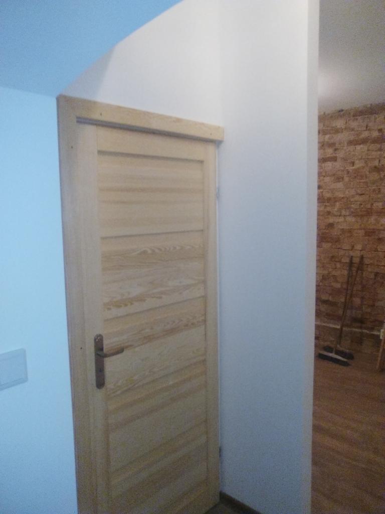 Surowe drewniane drzwi z poziomymi panelami w jasnym wnętrzu, widoczne fragmenty ceglanej ściany w tle. Klamka zamontowana.