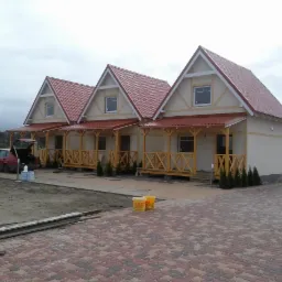 Trzy identyczne, nowo wybudowane domki letniskowe z czerwonymi dachami i drewnianymi werandami, widoczne z zewnątrz, z samochodem z otwartym bagażnikiem i wiaderkami z farbą w pobliżu.