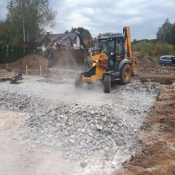 INŻYNIERIA PAWEŁ SKIBIŃSKI - Żółta koparko-ładowarka JCB rozprowadza kamień na przygotowanym gruncie pod budowę, ubijarka wibracyjna obok, w tle dom w budowie i zaparkowany samochód.