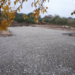 INŻYNIERIA PAWEŁ SKIBIŃSKI - Przygotowany teren pod budowę domu, wysypany żwirem i wyrównany, z widoczną koparką w tle i paletą materiałów budowlanych po lewej stronie, jesienne liście na pierwszym planie.