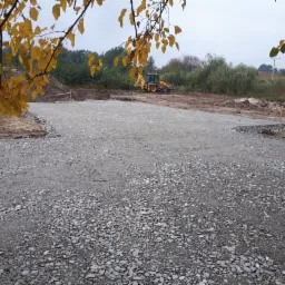 Przygotowany teren pod budowę domu, wysypany żwirem i wyrównany, z widoczną koparką w tle i paletą materiałów budowlanych po lewej stronie, jesienne liście na pierwszym planie.