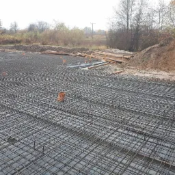 Zbrojenie płyty fundamentowej domu jednorodzinnego z widocznymi pionowymi prętami dystansowymi i rurami kanalizacyjnymi, przygotowane do zalania betonem, w tle hałda ziemi i drzewa.