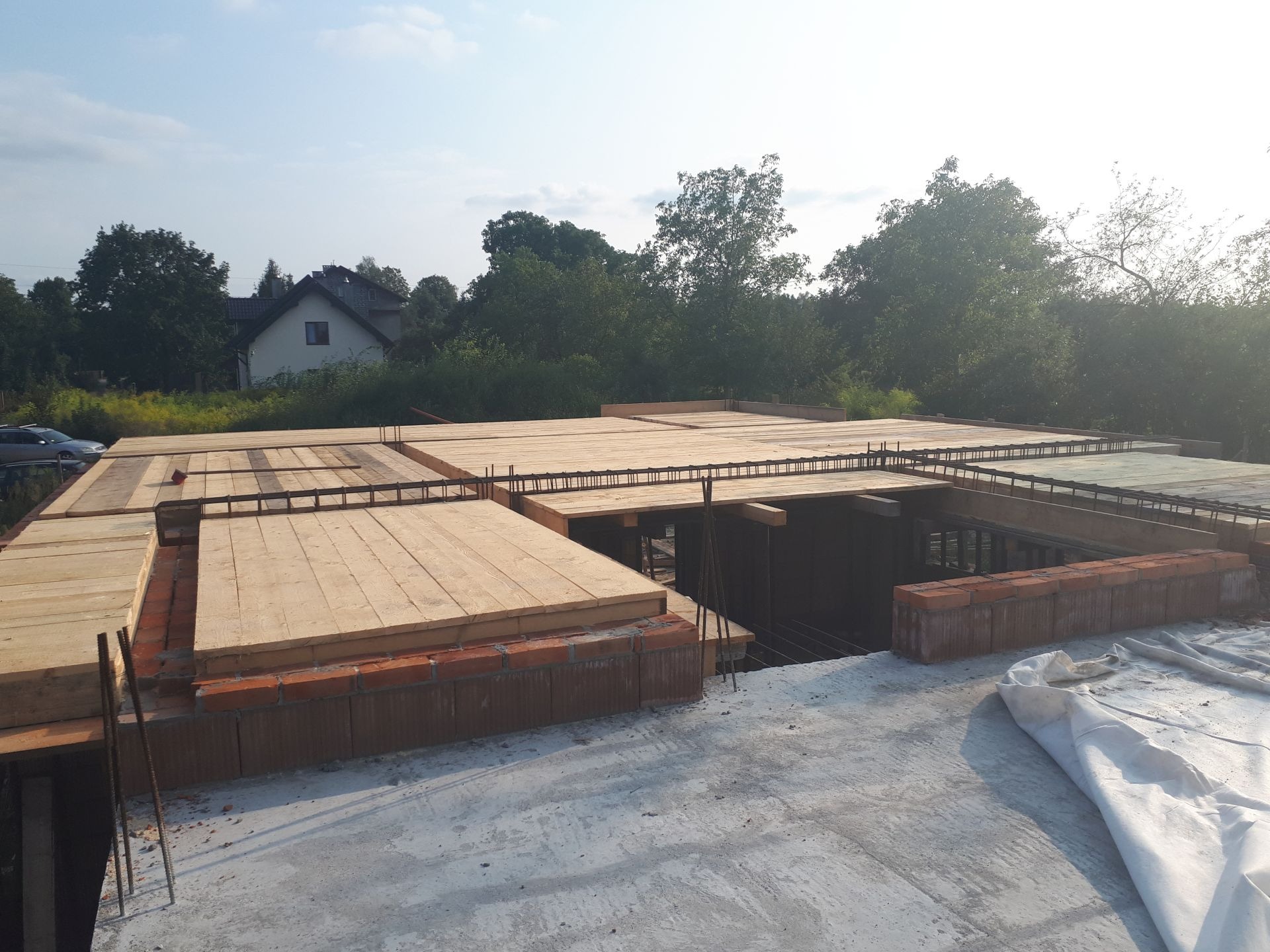 Wiązanie zbrojenia i układanie drewnianego szalunku na stropie budynku w trakcie budowy, widoczne cegły na obrzeżach, w tle zieleń i dom jednorodzinny.