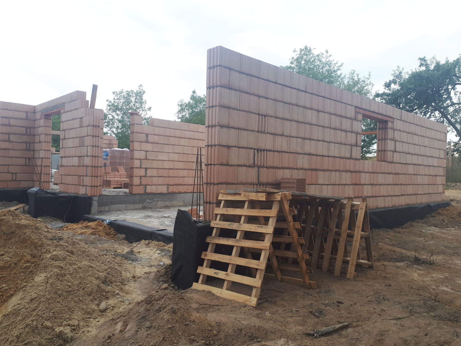 Wznoszenie ścian parteru z pustaków ceramicznych na fundamencie z izolacją przeciwwilgociową, widoczne drewniane rusztowanie i zapas materiału budowlanego.