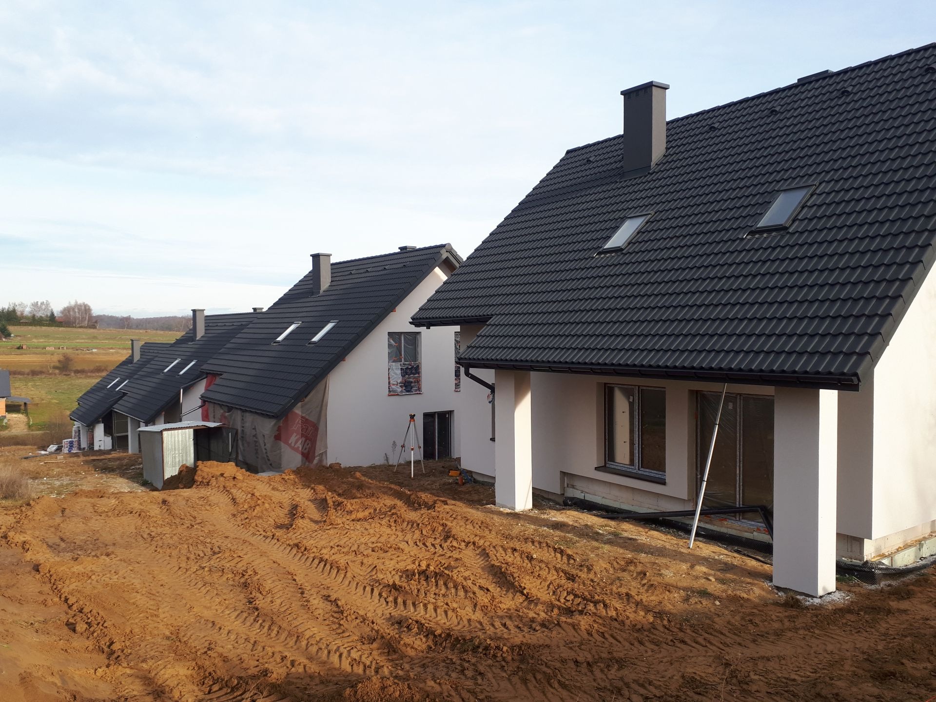 Wizualizacja szeregu domów jednorodzinnych w trakcie budowy, z czarną dachówką i białymi elewacjami, na nierównym terenie pokrytym ziemią, z widocznymi elementami konstrukcyjnymi i oknami bez...