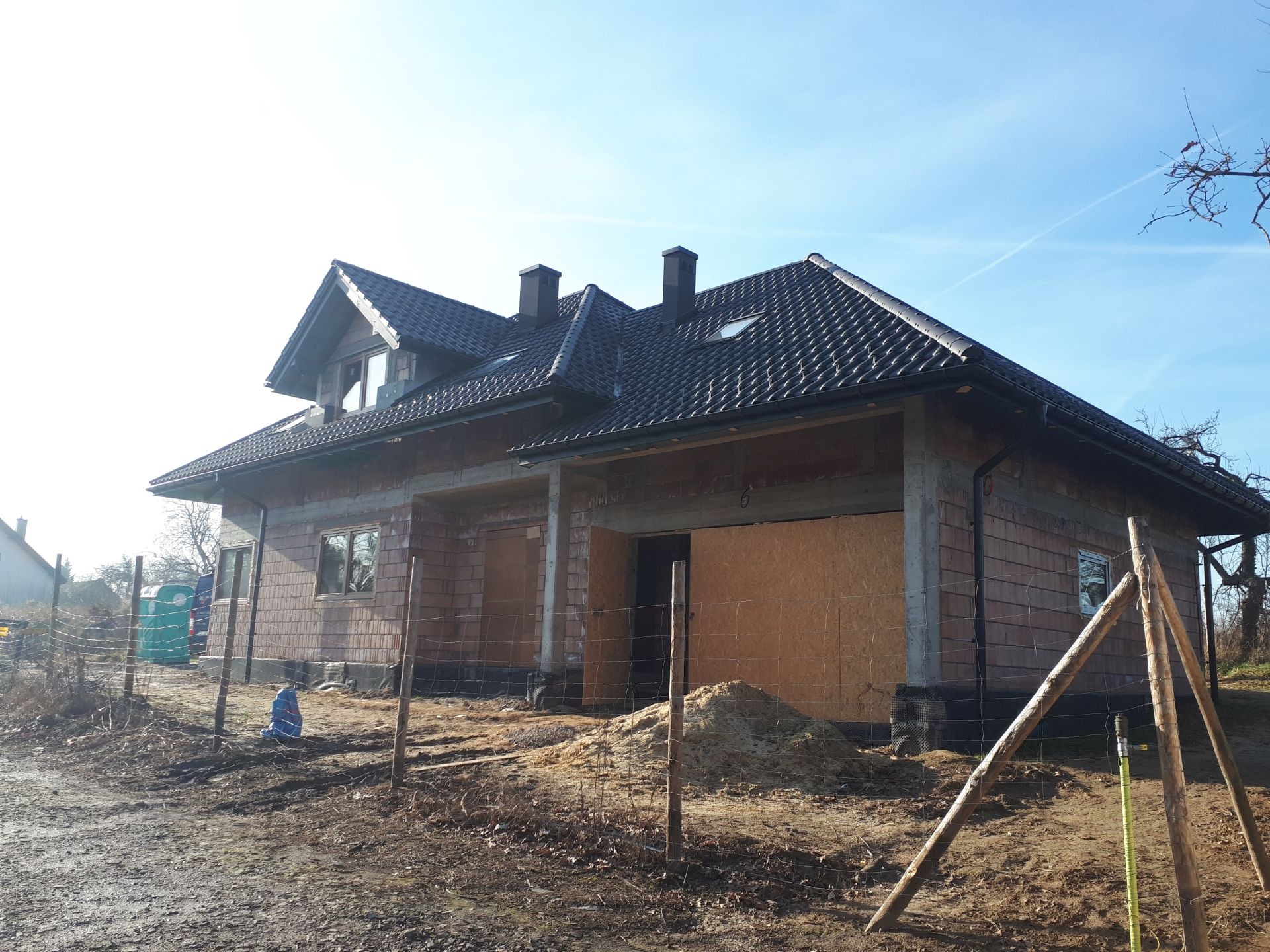 Surowy stan domu z czerwonej cegły z ciemną dachówką, widoczne okna, kominy, zabezpieczone otwory, teren budowy z ogrodzeniem i stertą piachu.