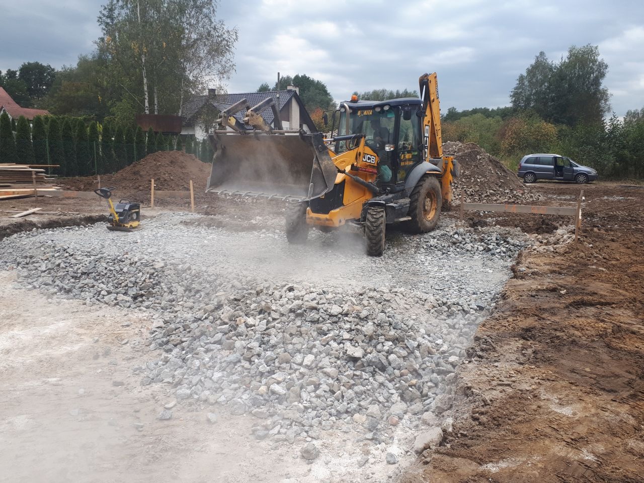 Żółta koparko-ładowarka JCB rozprowadza kamień na przygotowanym gruncie pod budowę, ubijarka wibracyjna obok, w tle dom w budowie i zaparkowany samochód.