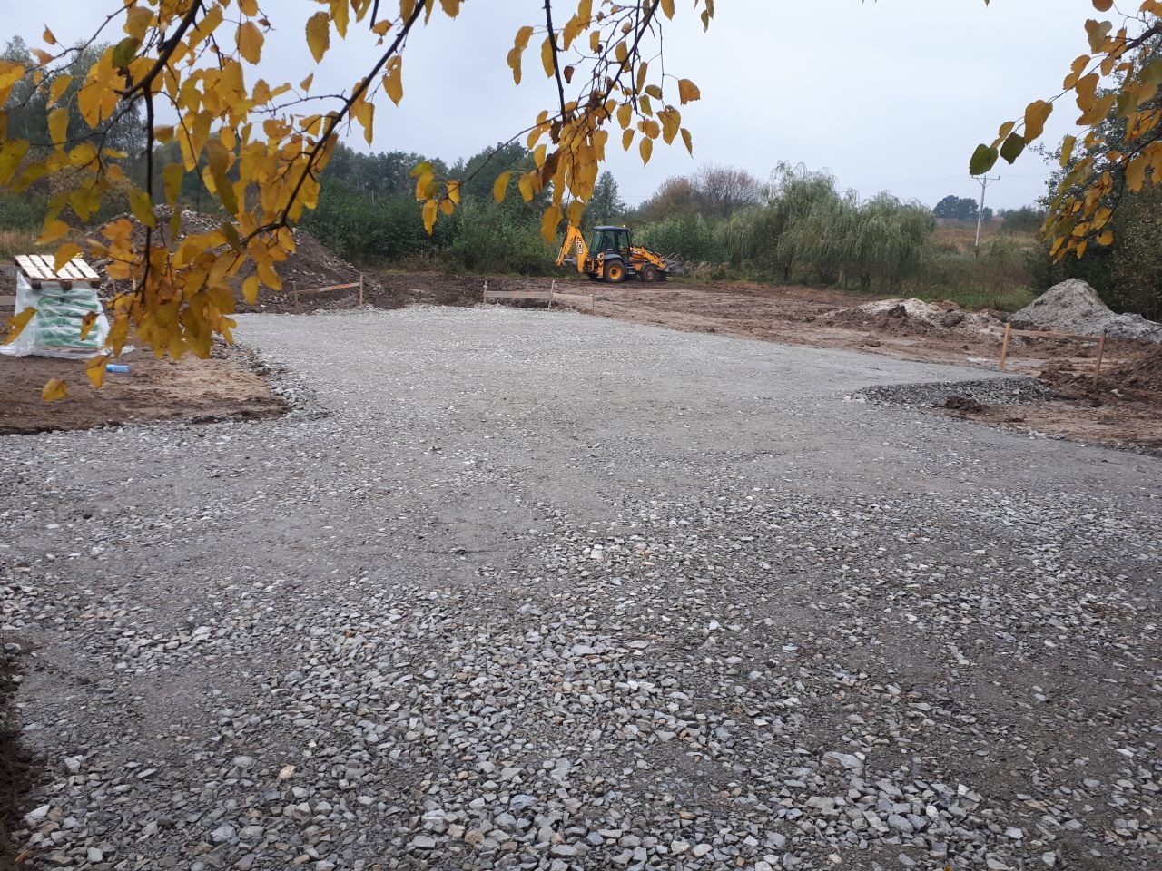 Przygotowany teren pod budowę domu, wysypany żwirem i wyrównany, z widoczną koparką w tle i paletą materiałów budowlanych po lewej stronie, jesienne liście na pierwszym planie.