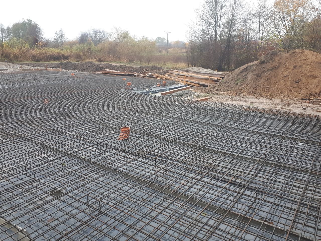 Zbrojenie płyty fundamentowej domu jednorodzinnego z widocznymi pionowymi prętami dystansowymi i rurami kanalizacyjnymi, przygotowane do zalania betonem, w tle hałda ziemi i drzewa.