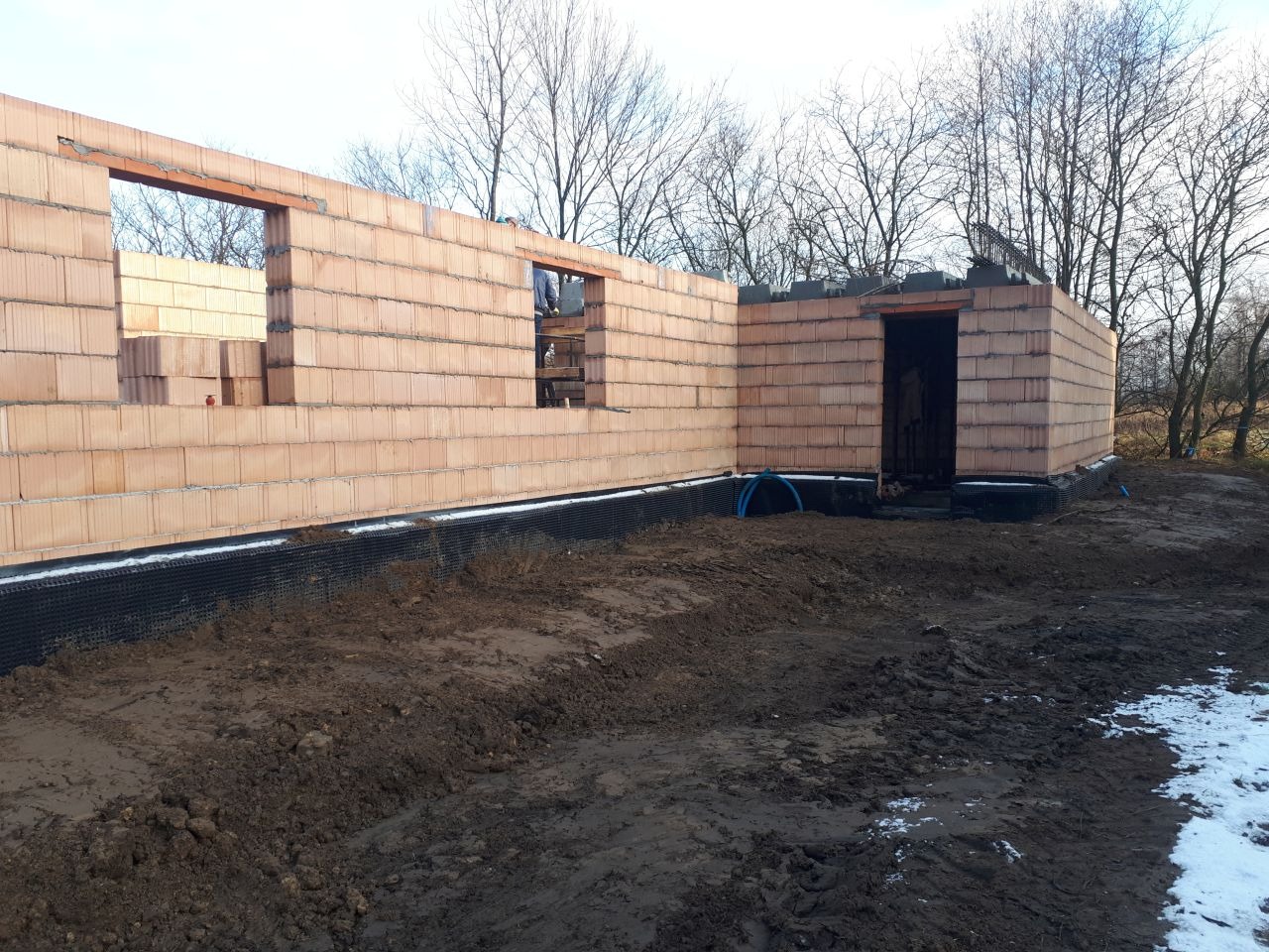 Wznoszenie ścian parteru domu z bloczków ceramicznych, widoczne otwory okienne i drzwiowe, czarna izolacja fundamentów, teren budowy z błotem i resztkami śniegu.
