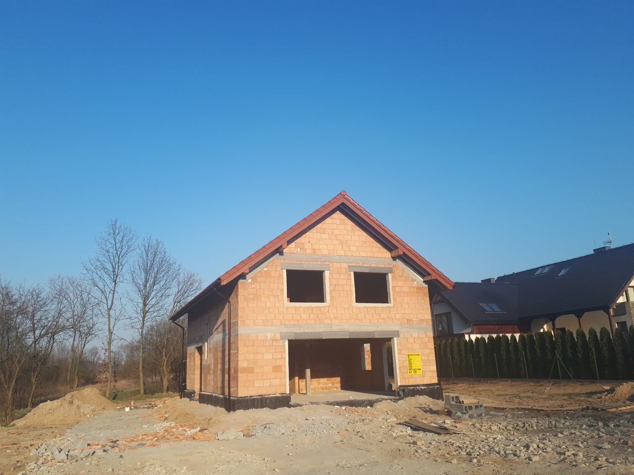 Surowy stan domu jednorodzinnego z czerwonej cegły, z widocznym garażem w bryle budynku, dach pokryty czerwoną dachówką, w tle błękitne niebo i sąsiednie domy.