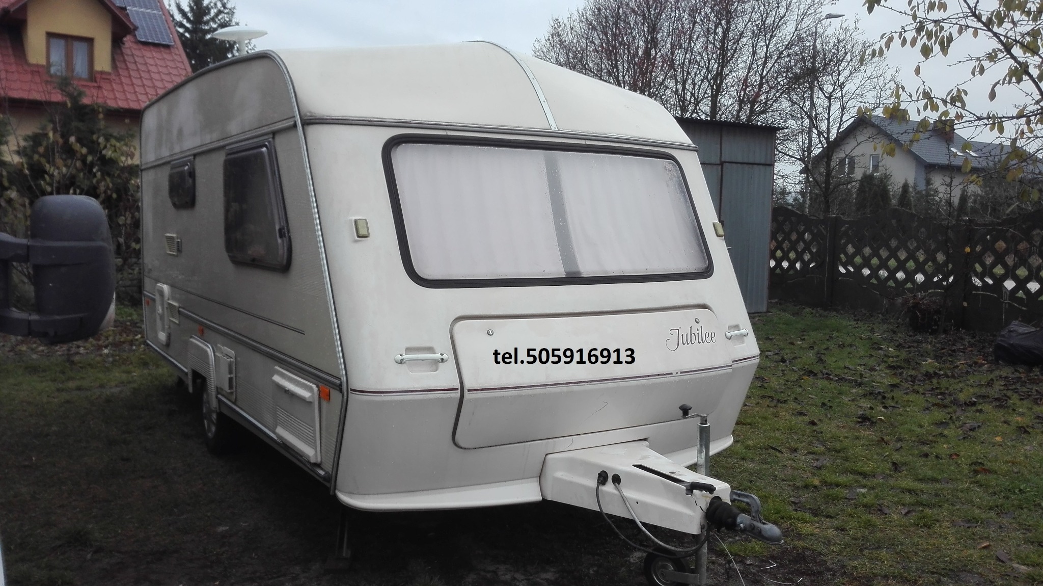 Używana przyczepa kempingowa Jubilee, widoczny numer telefonu, otoczenie: trawa, budynki mieszkalne i garaż.