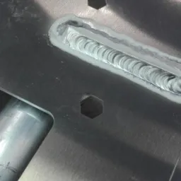 Detal spoiny na metalowej płycie z wyciętymi otworami pod klucz, widoczne charakterystyczne fale spawalnicze.