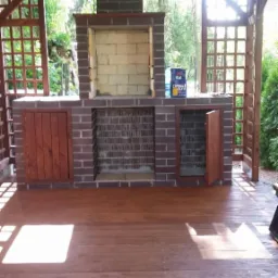 Drewniana altana z ceglanym grillem i kominem. Widoczne drzwi szafek pod grillem, podłoga z desek pomalowana bejcą. Na podłodze stoi puszka z farbą.