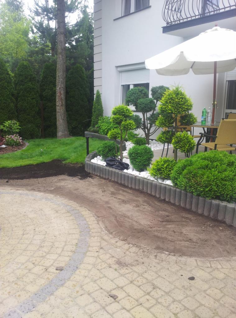 Ogród w trakcie realizacji: rabata z ozdobnymi krzewami i białymi kamieniami, obrzeże z betonowych elementów, kostka brukowa na podwórku, część trawnika i domu.