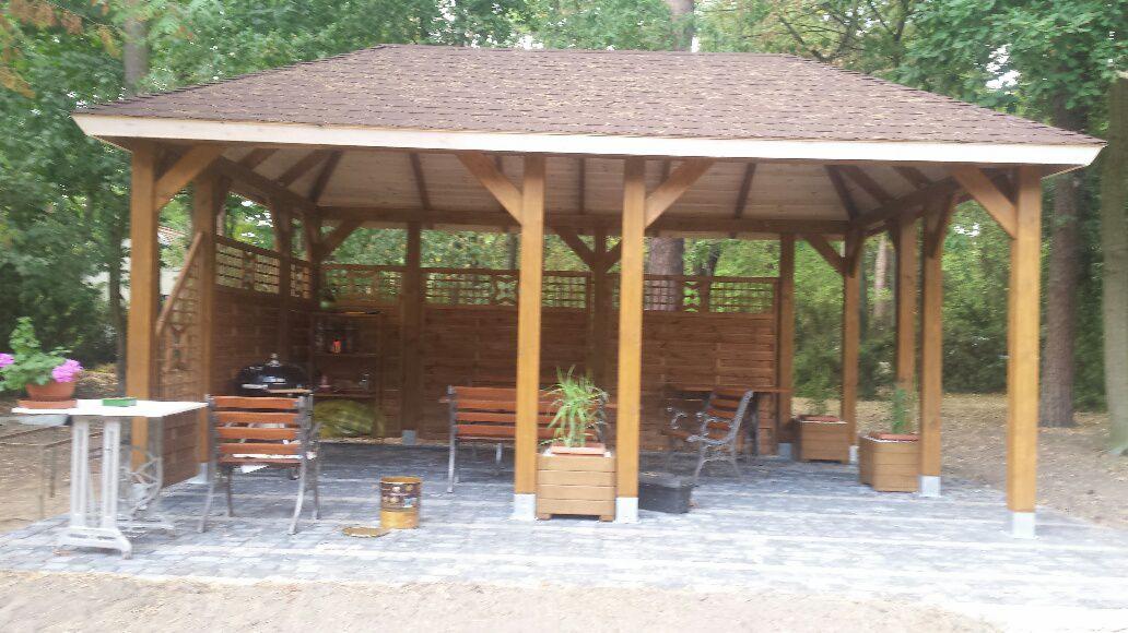 Drewniana altana ogrodowa z ażurowymi ścianami, wyposażona w meble i grill, otoczona zielenią. Podłoga wyłożona kostką brukową. Stół z kwiatami po lewej.