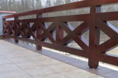 Ciemnobrązowa, drewniana balustrada tarasu z dekoracyjnym wzorem rombów, mocowana na jasnych płytkach. W tle widoczne drzewa bez liści.