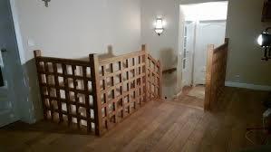 Drewniana barierka schodowa z kwadratową kratą, oświetlona lampą ścienną, na tle białych drzwi i podłogi z paneli. Widok z dołu schodów.