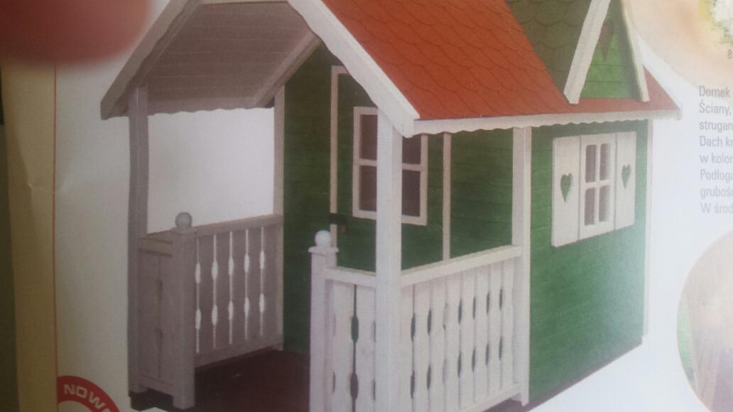Zielony domek zabaw dla dzieci z czerwonym dachem, białymi okiennicami z serduszkami i werandą. Elementy drewniane, widoczne detale konstrukcyjne i wykończeniowe.
