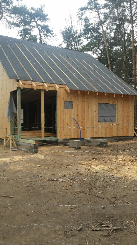 Budowa drewnianej altany w lesie: surowe ściany, częściowo pokryty dach papą, widoczne elementy konstrukcyjne, fundamenty z betonu. Prace w toku, otoczenie naturalne.