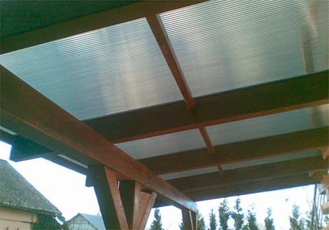 Drewniana konstrukcja z przezroczystym zadaszeniem, widok od dołu. Elementy konstrukcyjne z ciemnego drewna, zadaszenie z tworzywa sztucznego z widoczną fakturą. Na zewnątrz budynki i drzewa.