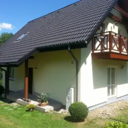 Ukończony dom jednorodzinny z ciemną dachówką, drewnianym balkonem i jasną elewacją w słoneczny dzień. Widoczne rośliny i elementy ogrodu.