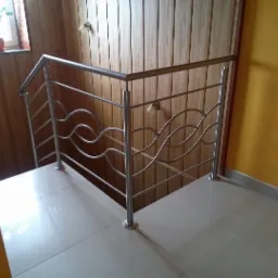 Barierka schodowa ze stali nierdzewnej z poziomymi prętami w kształcie fal, zamontowana na białych płytkach podłogowych, przy ścianie wyłożonej boazerią i żółtej ścianie.