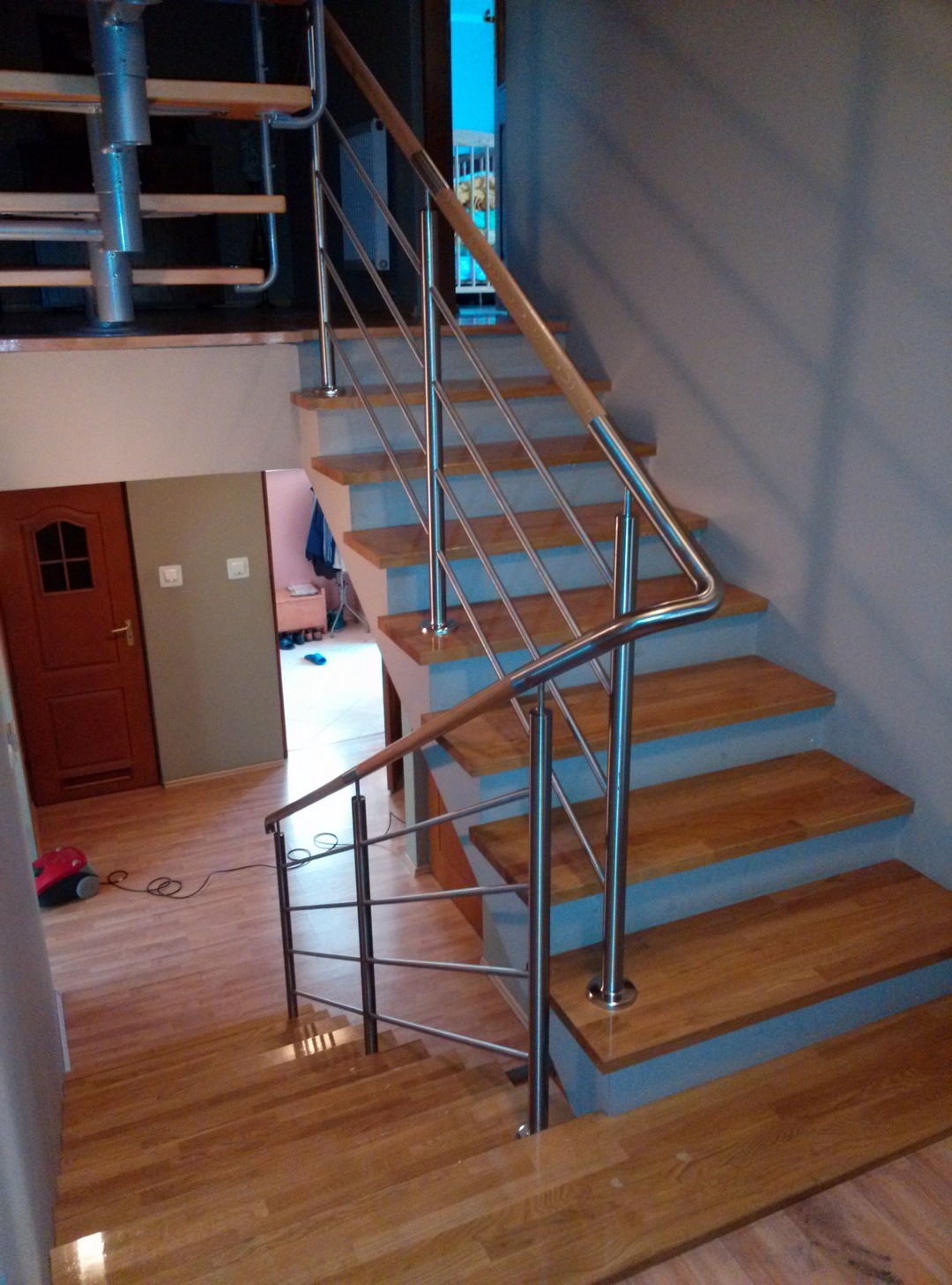Wewnętrzne schody z drewnianymi stopniami i balustradą z elementami ze stali nierdzewnej oraz drewnianą poręczą, widoczne z dołu schodów, z widokiem na podłogę z paneli i otwarte drzwi do kolejnego...