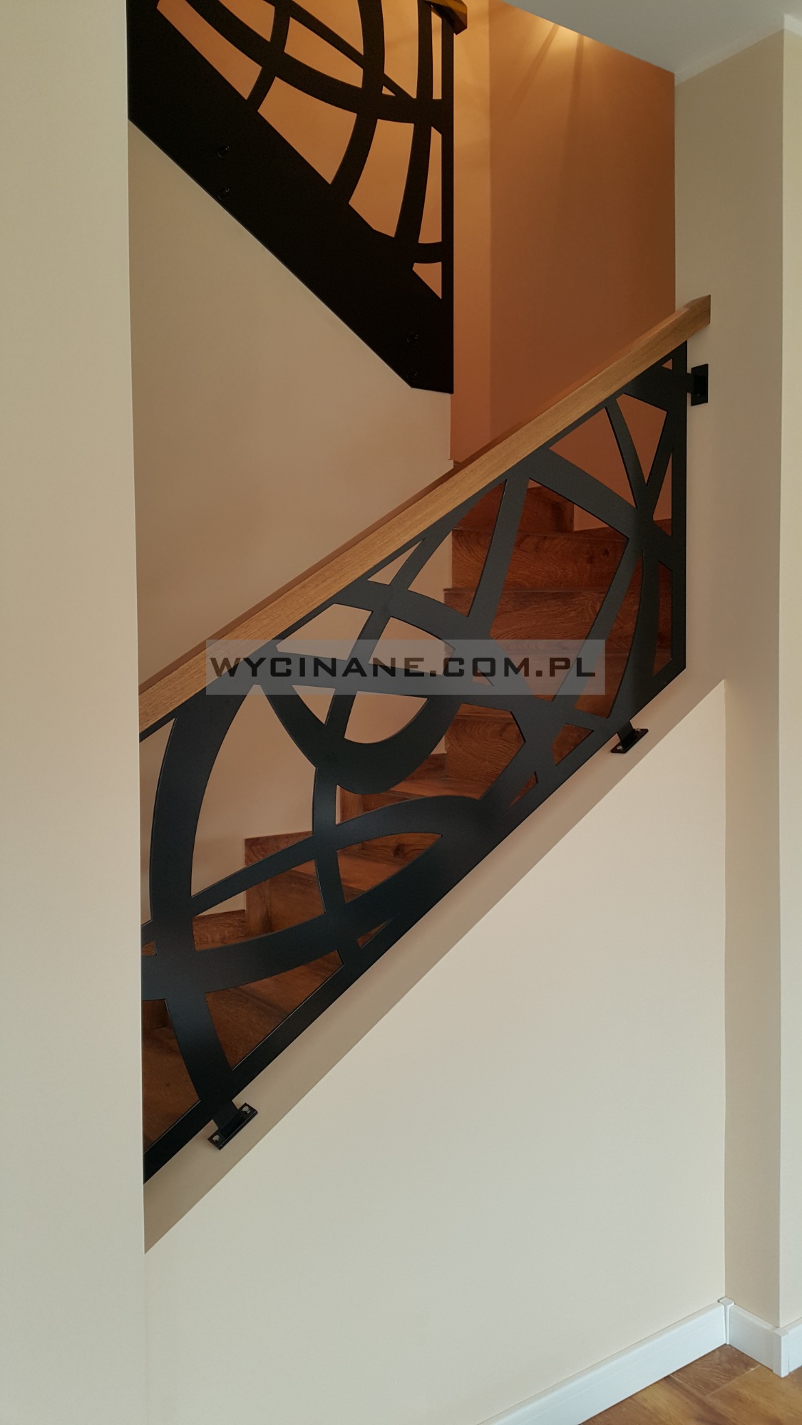 Schody drewniane z nowoczesną, czarną, metalową balustradą o abstrakcyjnym wzorze i drewnianą poręczą, widok z dołu.