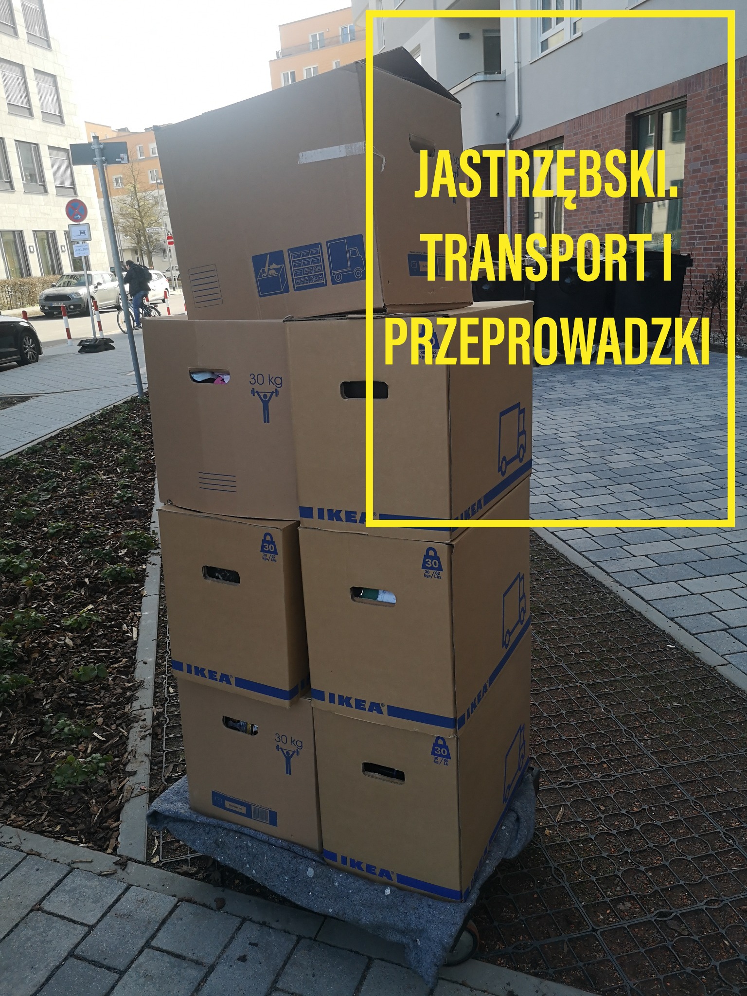 Stos kartonowych pudeł IKEA, oznaczonych jako 30 kg, ustawionych na wózku transportowym, przed budynkiem mieszkalnym.