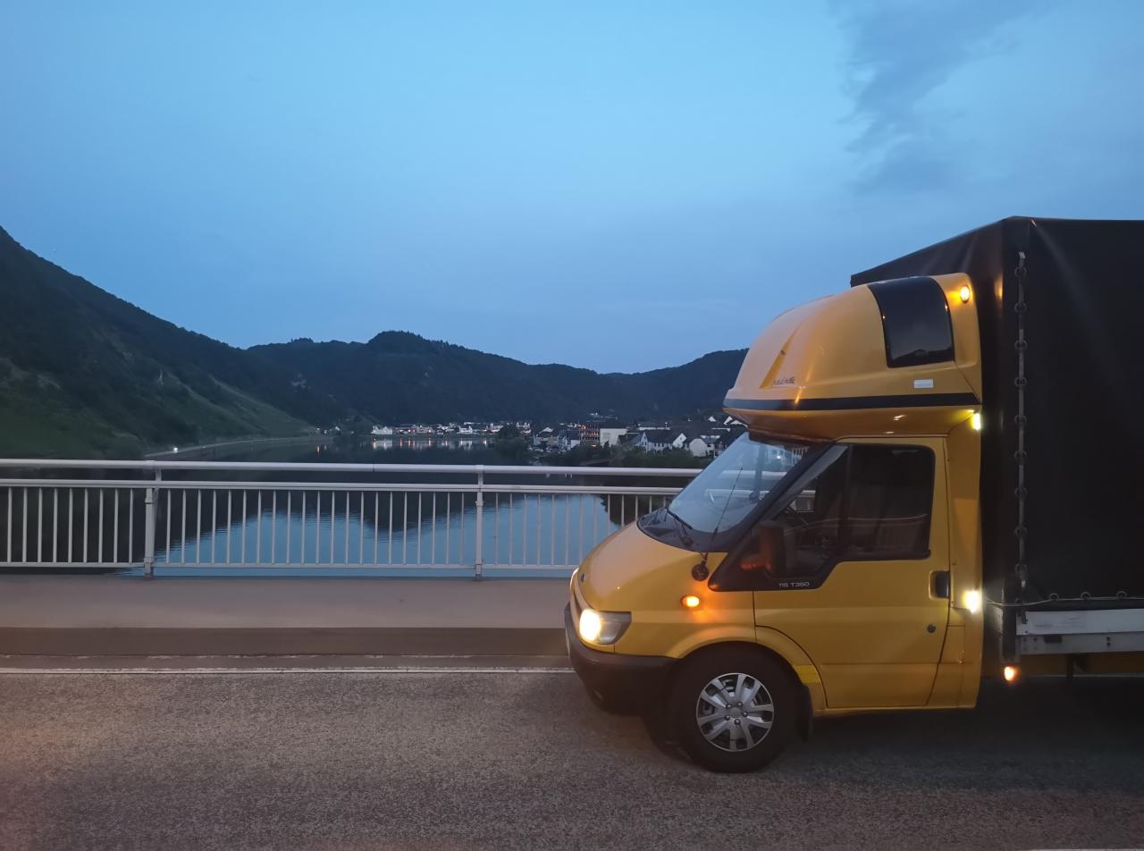Żółty bus transportowy z plandeką parkuje na moście; w tle widoczne góry i miasteczko w wieczornym oświetleniu. Ujęcie z boku, światła pojazdu włączone.