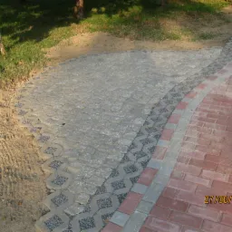 Ścieżka z kostki brukowej o różnej fakturze i kolorystyce, obrzeże z ażurowych elementów wypełnionych grysem, trawnik w tle. Słoneczny dzień, widok z góry.