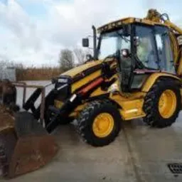 Żółto-czarna koparko-ładowarka Caterpillar z łyżką z zębami na betonowej nawierzchni. W tle błękitne kontenery i drzewa pod pochmurnym niebem.