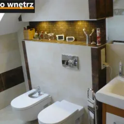 Jasna łazienka na poddaszu z beżowymi płytkami, brązowym pasem płytek i mozaiką nad blatem. Umywalka, bidet i toaleta w minimalistycznym stylu.