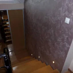 Schody drewniane z oświetleniem punktowym, ściana z dekoracyjnym tynkiem w odcieniach brązu i beżu, widok z góry na dół, fragment balustrady.