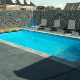 Błękitny basen otoczony szarą kostką, z gabionową ścianą w tle i czterema leżakami, tworzy nowoczesną strefę relaksu w ogrodzie.