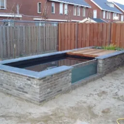 Nowoczesne oczko wodne z ceglaną obudową, częściowo przykryte drewnianym podestem, z widoczną szklaną ścianą i roślinnością. Tło stanowi płot i zabudowa mieszkalna.