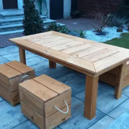 Drewniany stół z ławką i dwoma pufami na tarasie z szarej kostki, w tle trawnik i roślinność. Widoczne detale: liny jako uchwyty puf.