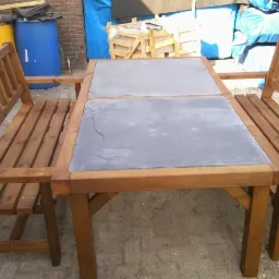 Drewniany stół ogrodowy z ławkami i blatem z szarego kamienia na zewnątrz, z niebieskimi beczkami i skrzynią w tle.