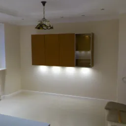 Nowoczesna aranżacja wnętrza z podświetlanymi szafkami wiszącymi w odcieniach brązu i beżu, oświetlonymi od dołu. Widoczne krzesła i lampa wisząca.