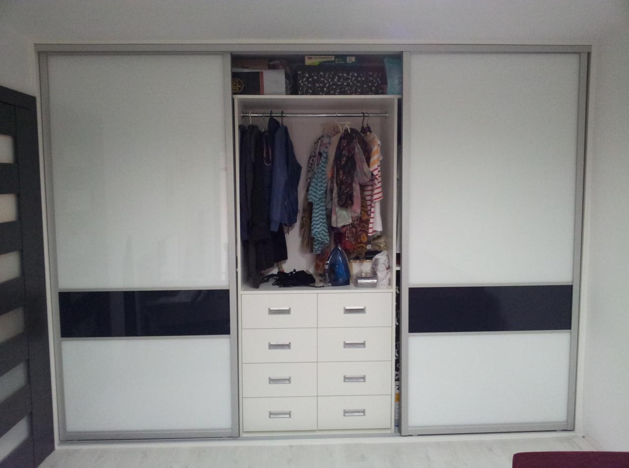 Biała szafa wnękowa z przesuwnymi drzwiami, z czarnym pasem na środku. Widoczna zawartość szafy: ubrania na wieszakach i komoda z szufladami. Minimalistyczny design.