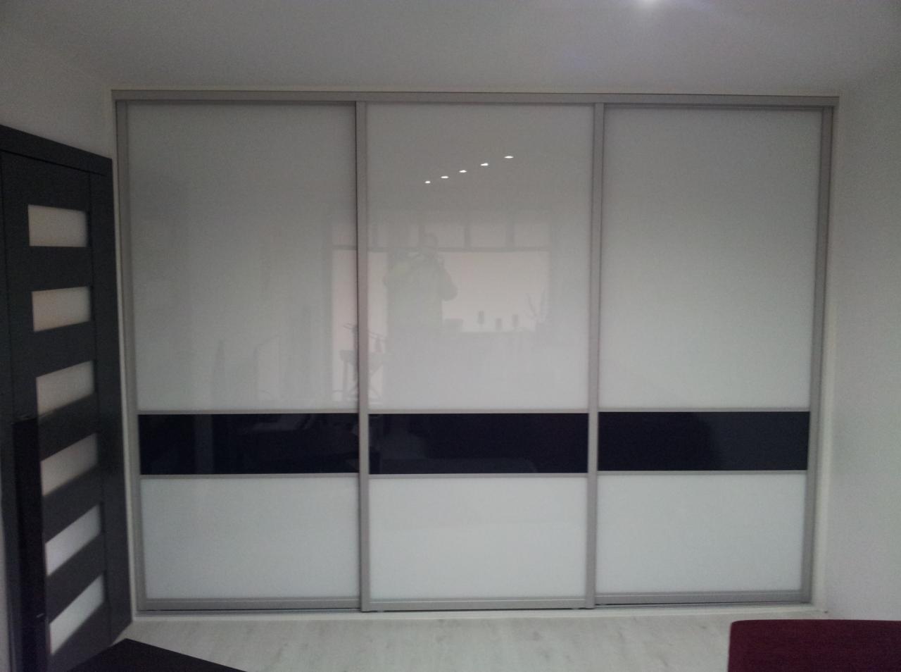 Biała szafa wnękowa z przesuwnymi drzwiami i czarnym pasem pośrodku, widoczna obok ciemna rama drzwi.