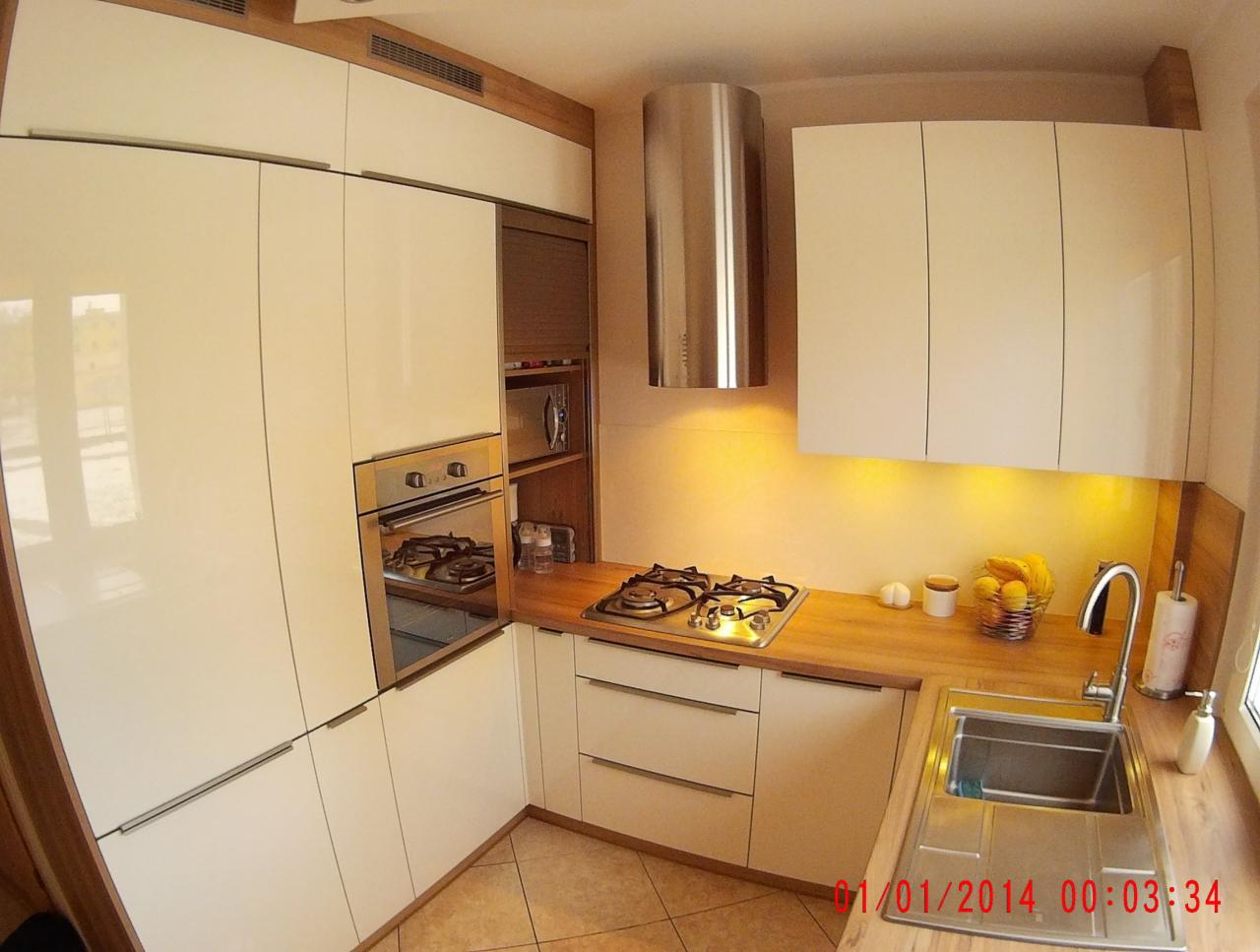 Biała kuchnia z drewnianymi akcentami. Widoczny zlew, płyta gazowa, piekarnik i szafki. Uwagę zwraca cylindryczny okap i oświetlenie podszafkowe.