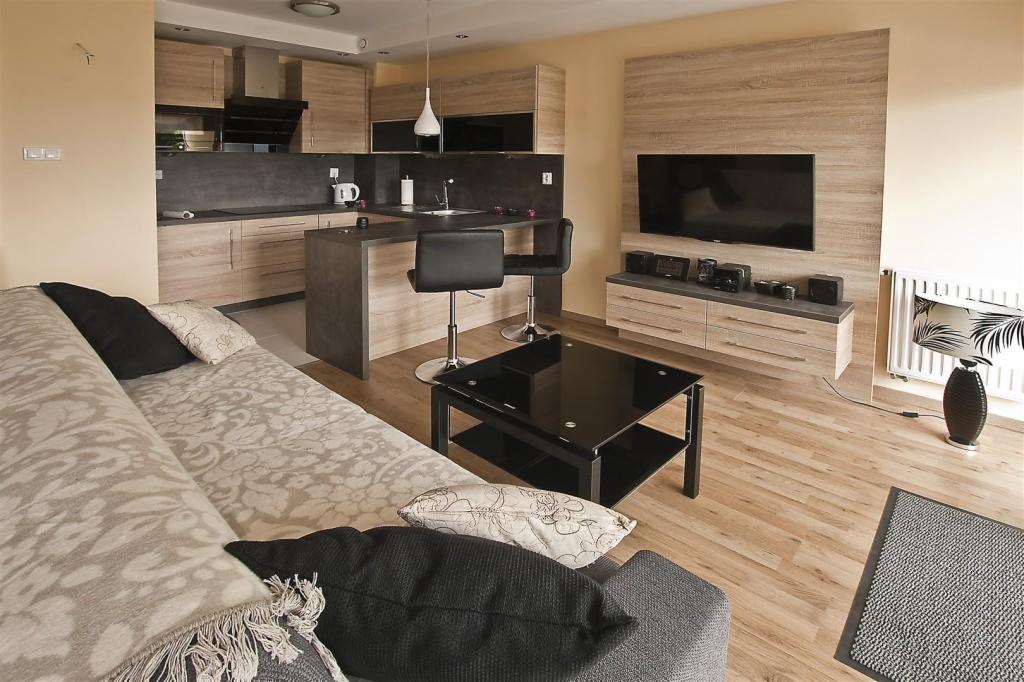 Jasne wnętrze z drewnianymi meblami: kuchnia otwarta na salon. Wyspa kuchenna z hokerami, kanapa z poduszkami, czarny stolik kawowy i szafka RTV z telewizorem.