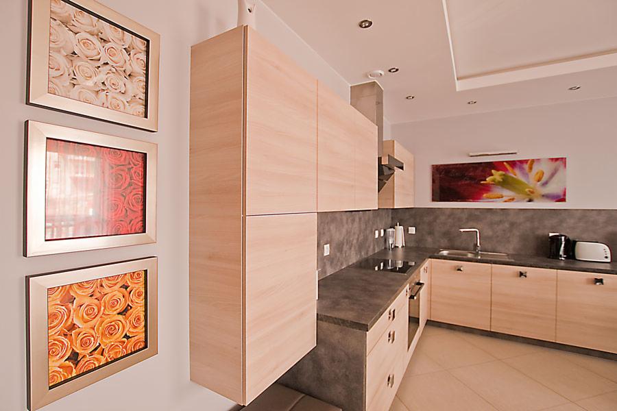 Jasna, drewniana zabudowa kuchenna z szarym blatem w Pile. Na ścianie trzy obrazy z motywem róż. Nowoczesne oświetlenie sufitowe, minimalistyczny design.