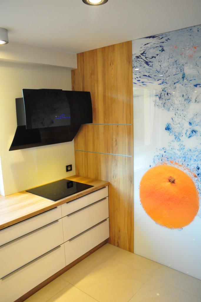 Nowoczesna kuchnia z białymi szafkami, drewnianym blatem i panelem ze zdjęciem pomarańczy. Czarny okap nad płytą indukcyjną. Minimalistyczny design.