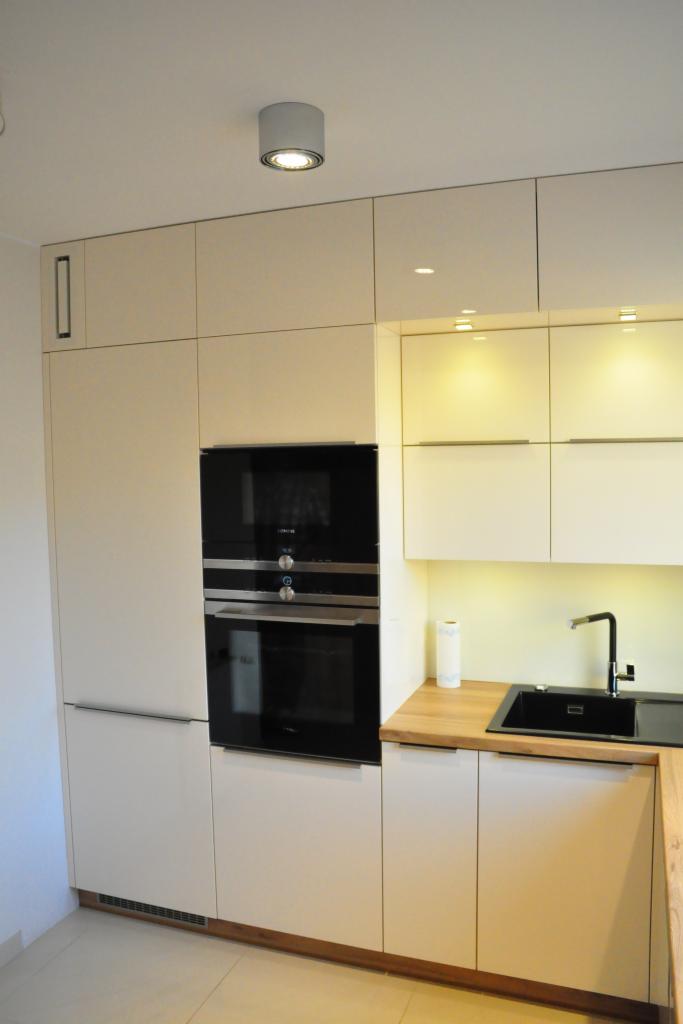 Nowoczesna, jasna kuchnia na wymiar z zabudowaną czarną piekarnikiem i mikrofalówką, drewnianym blatem i czarnym zlewem. Minimalistyczny design.