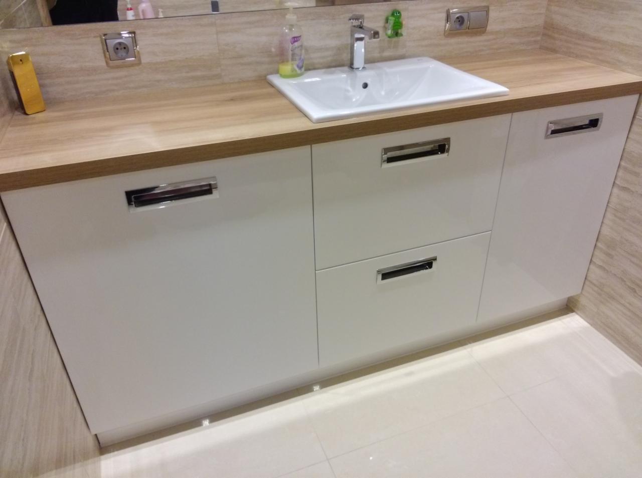 Biała szafka łazienkowa z drewnianym blatem, chromowanymi uchwytami i umywalką. Widoczne gniazdka elektryczne i elementy dekoracyjne na blacie. Jasne płytki podłogowe.