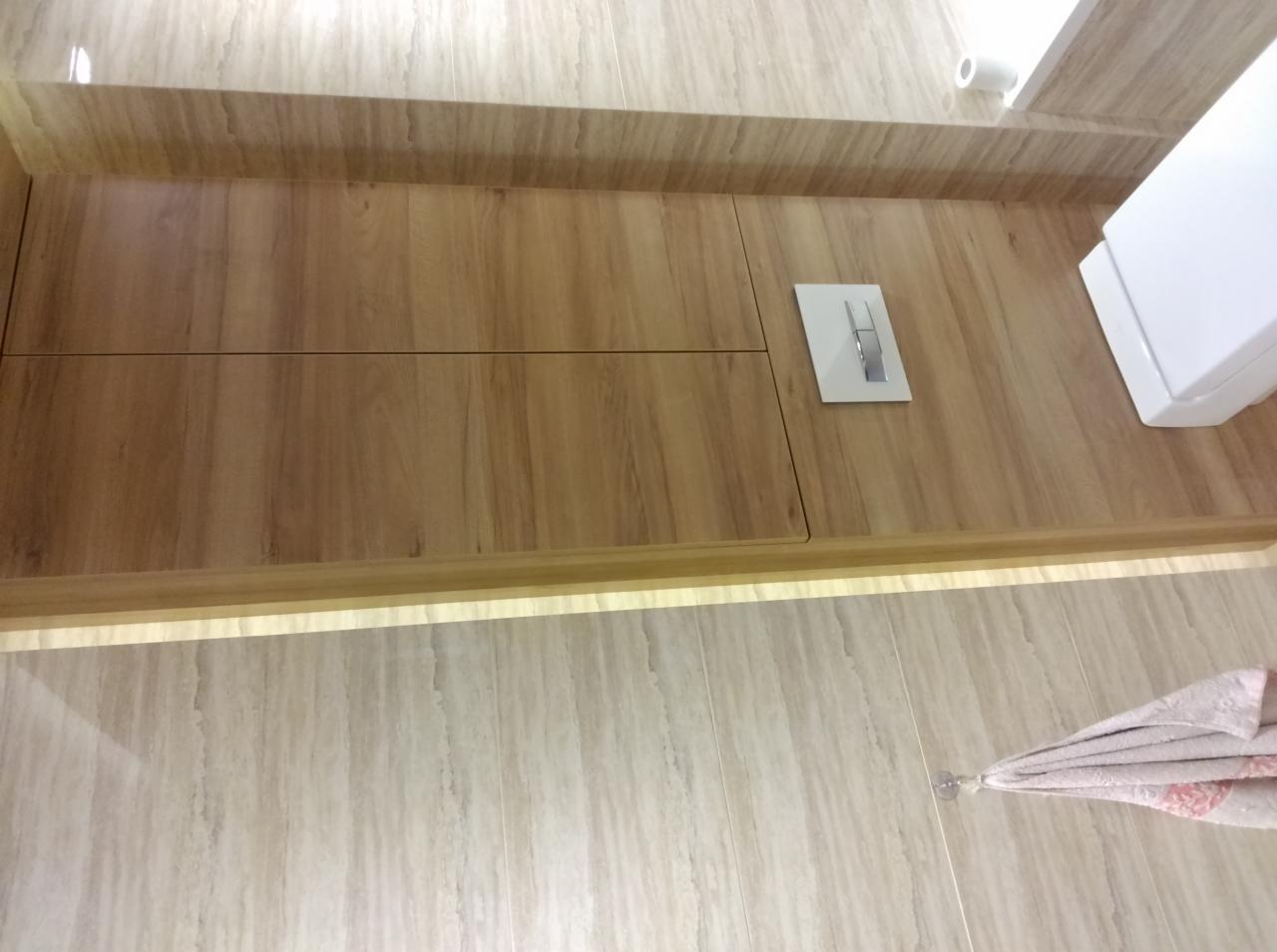 Drewniana zabudowa toalety z ukrytym systemem spłukiwania i oświetleniem LED. Całość w jasnych, beżowych odcieniach, z widoczną armaturą i fragmentem toalety.