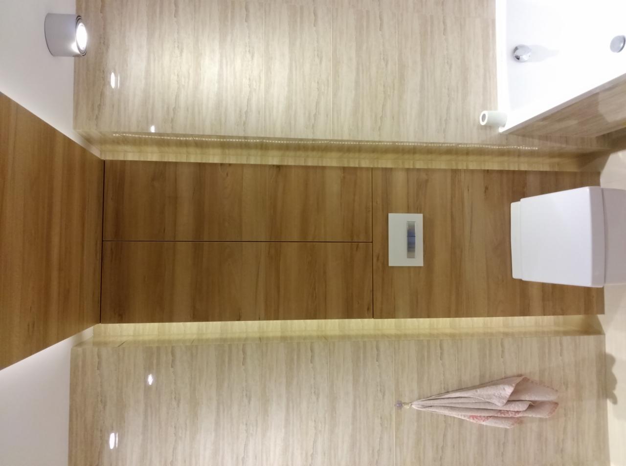 Nowoczesna łazienka z drewnianymi szafkami w zabudowie, podświetleniem LED i beżowymi płytkami. Widoczna umywalka, toaleta i ręcznik.